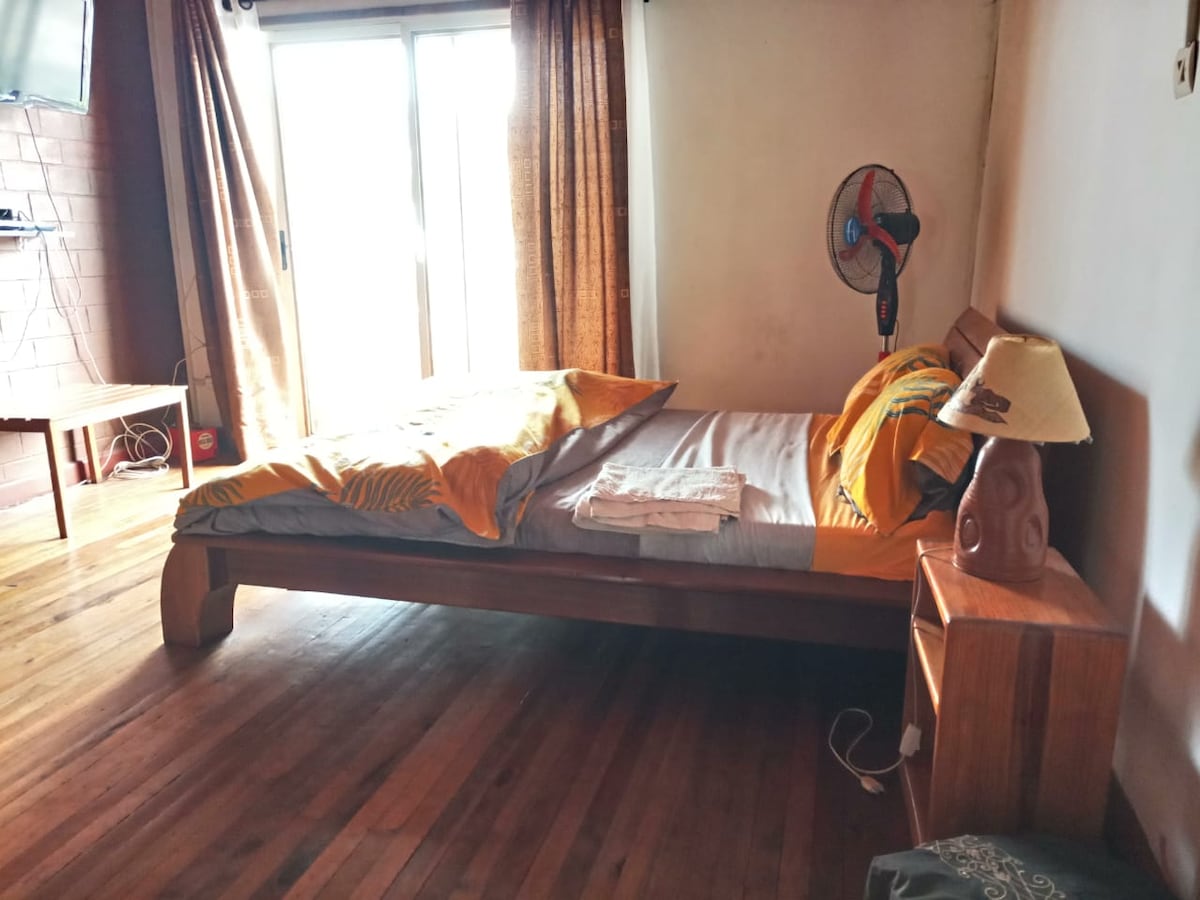 A cozy sleeping area is presented with a wooden bed featuring neatly arranged blankets and a towel. Natural light enters through glass doors, illuminating the space. A small lamp and a fan are positioned beside the bed. Dark curtains frame the windows, enhancing the warm ambiance.