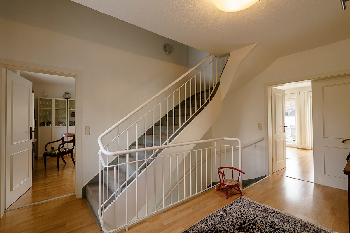 A spacious entryway is visible, featuring a wide staircase with a white railing leading to the upper floor. Light hardwood floors complement the neutral walls, and an area rug adds warmth. Doors to adjoining rooms are open, suggesting a welcoming atmosphere.
