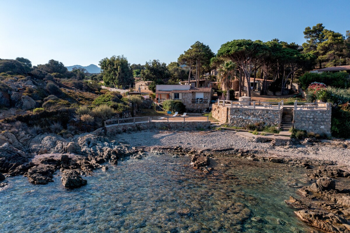 The image reveals a serene coastal property surrounded by lush greenery, with a small sandy beach leading to clear blue waters. Stone structures are visible, along with natural rock formations lining the shore. A gentle slope of terrain leads to a distant forested area.