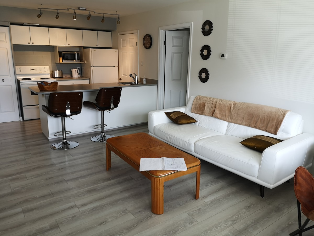 A modern living area features a white sofa with brown cushions, complemented by a wooden coffee table. In the background, a kitchen with bar seating and stainless-steel appliances is visible. Natural light floods the space, highlighting the light-colored flooring and neutral walls.