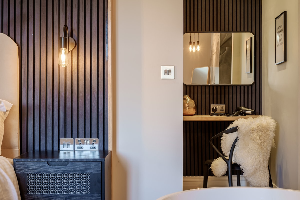 A modern bedroom space showcases a textured black accent wall behind a spacious bed. A stylish side table features practical charging outlets. Adjacent, a well-lit vanity area with a mirror and a chair is highlighted, alongside a luxurious standalone bathtub.