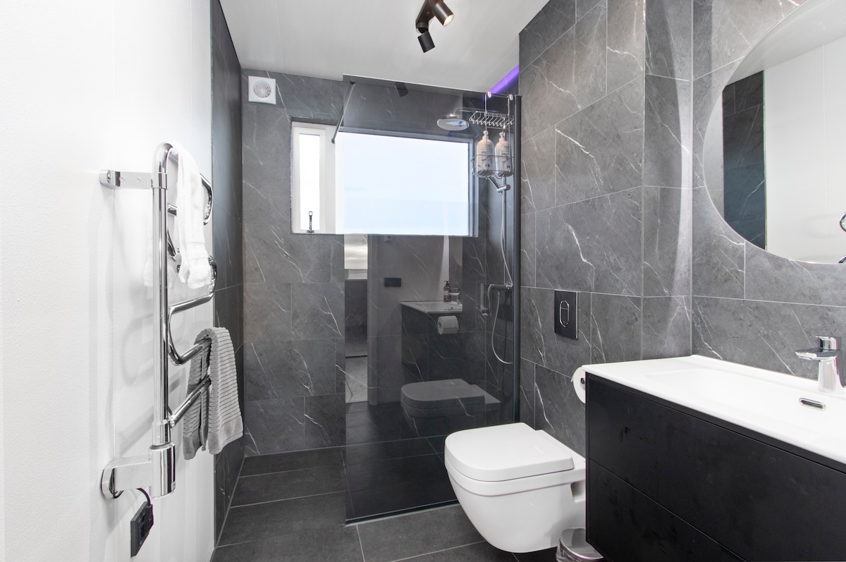 A modern bathroom is showcased, featuring sleek dark tiles and a glass shower enclosure. A wall-mounted towel rack with towels is visible alongside a minimalist vanity with a sink. Natural light is allowed in through a window, enhancing the spacious feel of the area.
