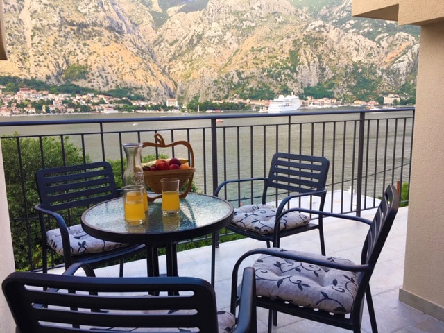 A terrace setting includes a glass table surrounded by four chairs, featuring a bowl of apples and two glasses. The backdrop presents a sweeping view of Kotor's mountainous landscape and waterfront, providing a serene atmosphere for relaxation.
