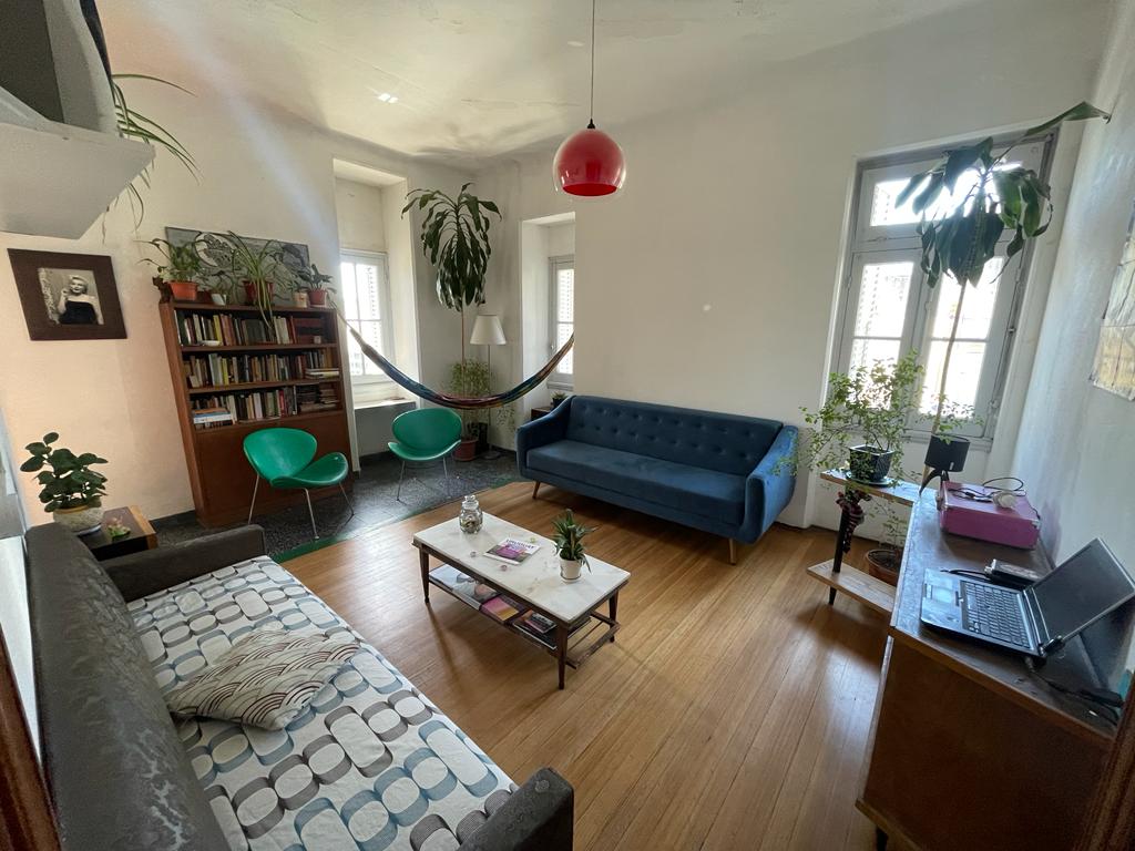 A spacious living area features a mix of seating options, including a blue couch, a patterned sofa, and green chairs. A coffee table sits in the center, surrounded by lush plants. Natural light streams in through large windows, enhancing the inviting ambiance.