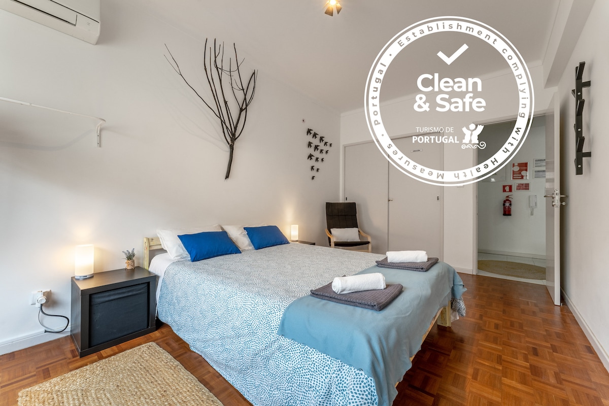A spacious bedroom features a comfortable double bed with crisp linens and two towels neatly arranged. A soft blue blanket covers the bed, while a decorative wall tree adds a touch of elegance. Natural light fills the room, creating a serene environment.