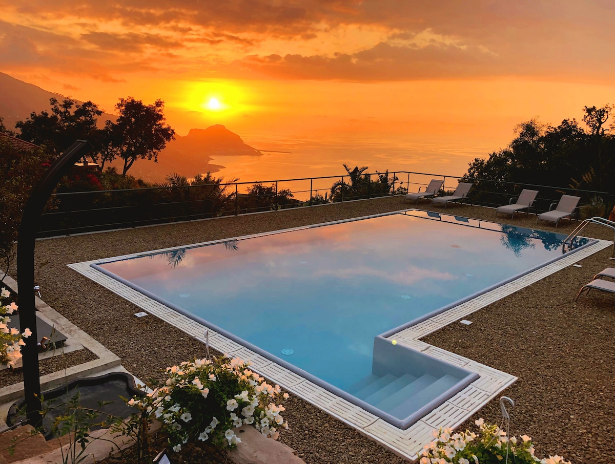 A serene infinity pool reflects the warm hues of a sunset over the Mediterranean Sea, surrounded by a beautifully landscaped area. Comfortable sun loungers are positioned nearby, offering a relaxing spot to enjoy the tranquil atmosphere and stunning views.