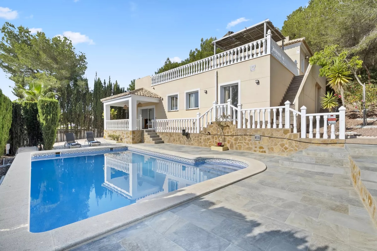 The villa is set against a backdrop of lush greenery, featuring a clear blue pool bordered by stone. A spacious terrace leads to the entrance, with steps ascending to the upper level. Palm trees and decorative plants add to the tranquil environment.
