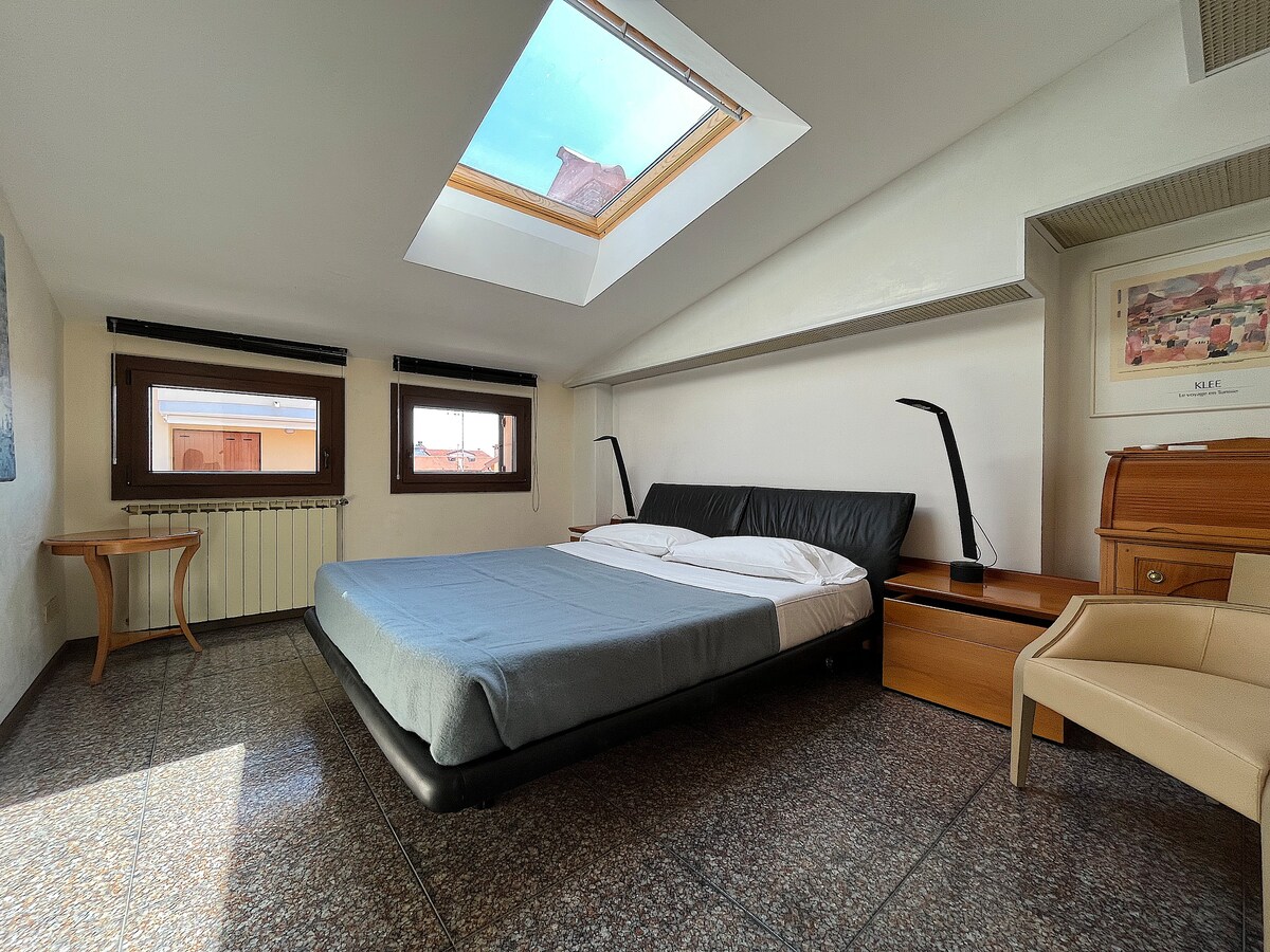 A well-lit bedroom features a spacious bed with a simple blue blanket. A skylight above allows natural light to brighten the room. Two windows on opposite walls provide additional views. Minimalist furniture includes a bedside table and a wooden dresser.