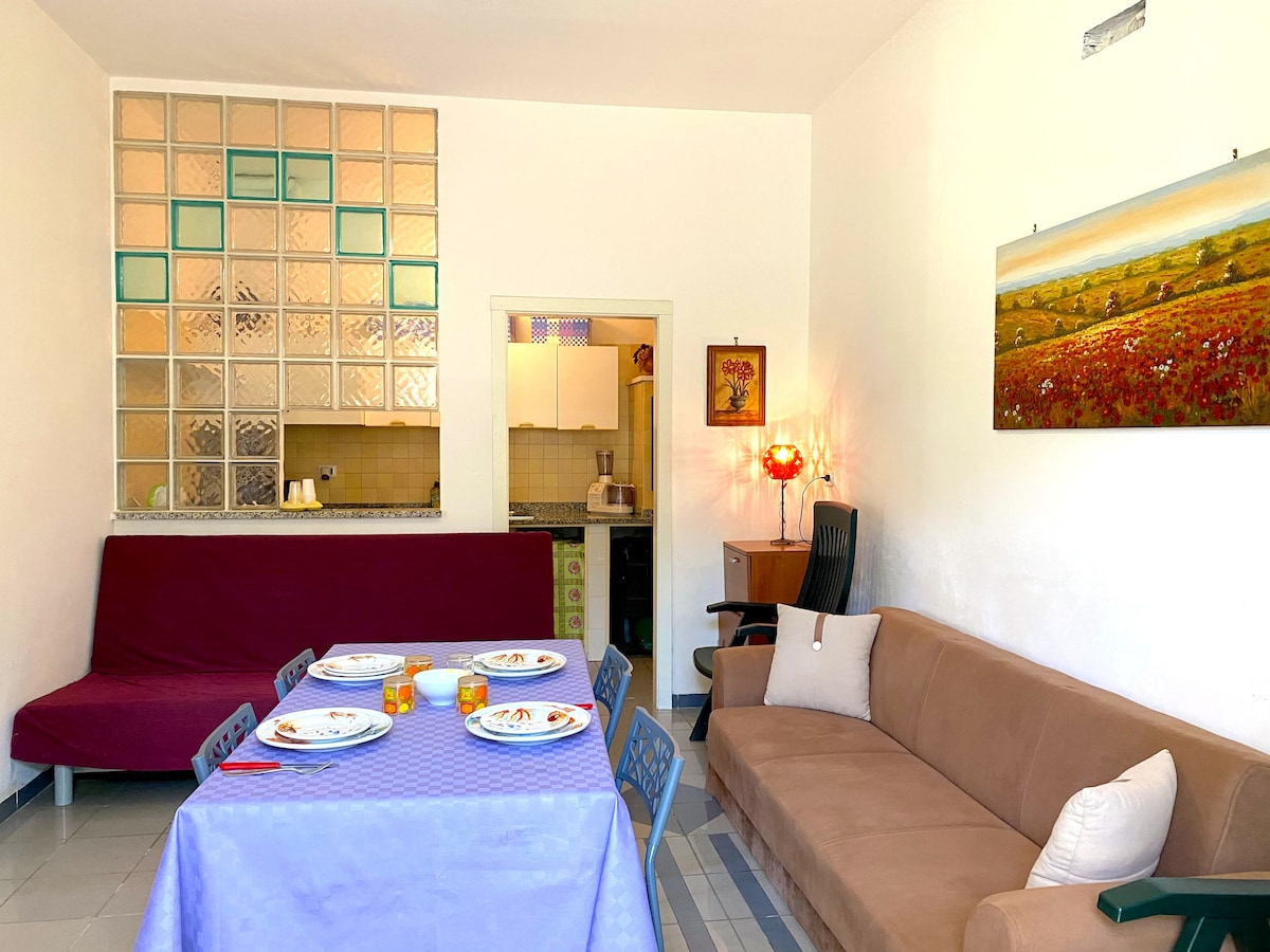 A spacious dining area features a blue tablecloth over a large rectangular table set for dinner. A brown sofa is positioned against one wall, while a workspace with a chair is visible in the background. Light filters through glass block windows, adding brightness to the room.