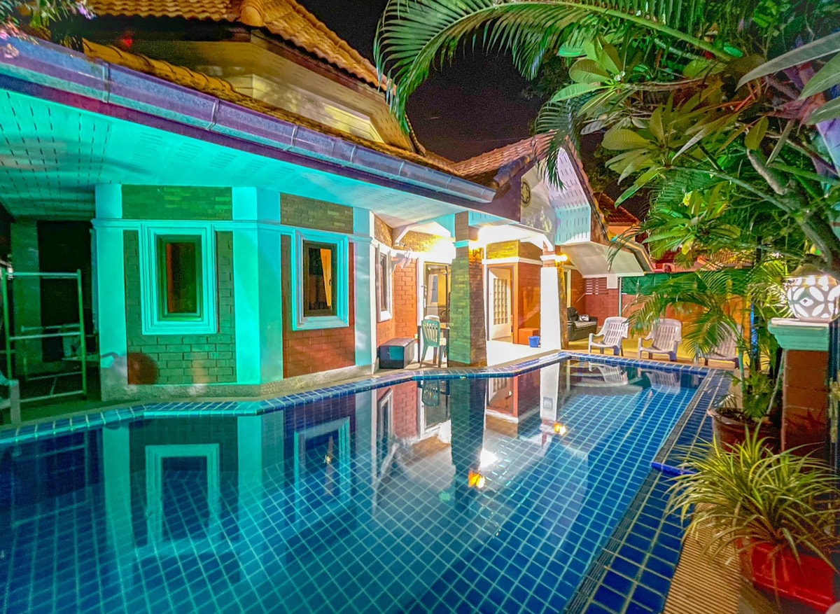 The image showcases a serene outdoor area featuring a private pool, surrounded by lush greenery and palm plants. The villa's exterior is illuminated with soft lighting, highlighting the architectural details. Comfortable seating options are visible near the pool, inviting relaxation.