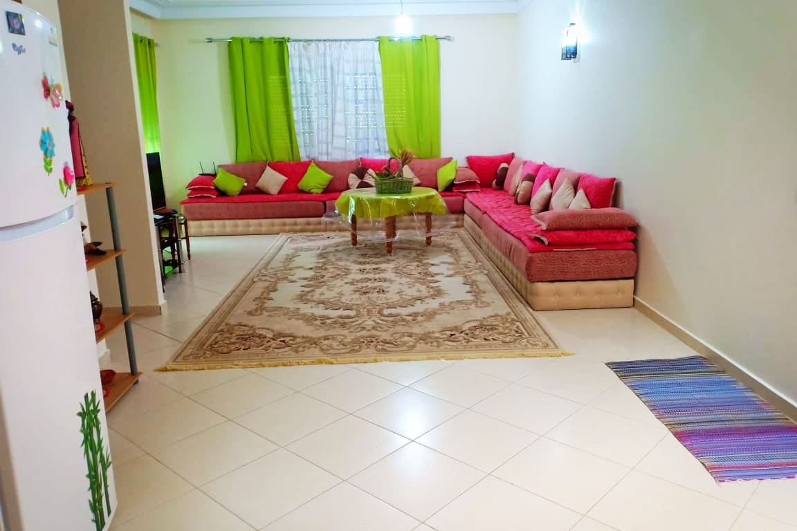 A spacious living area features a large, L-shaped sofa adorned with colorful cushions. A central table sits on an ornate area rug, creating a welcoming atmosphere. Sheer curtains in light colors allow natural light to fill the space, while decorative touches enhance the comfortable setting.