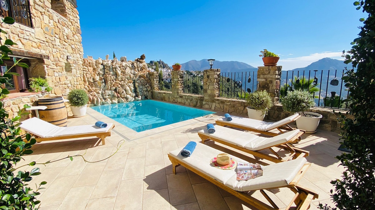 A tranquil outdoor area features a sparkling pool surrounded by a stone wall and mountain views. Four sun loungers with rolled towels sit adjacent to the water. Potted plants add greenery, while a barrel and a small table enhance the rustic vibe.