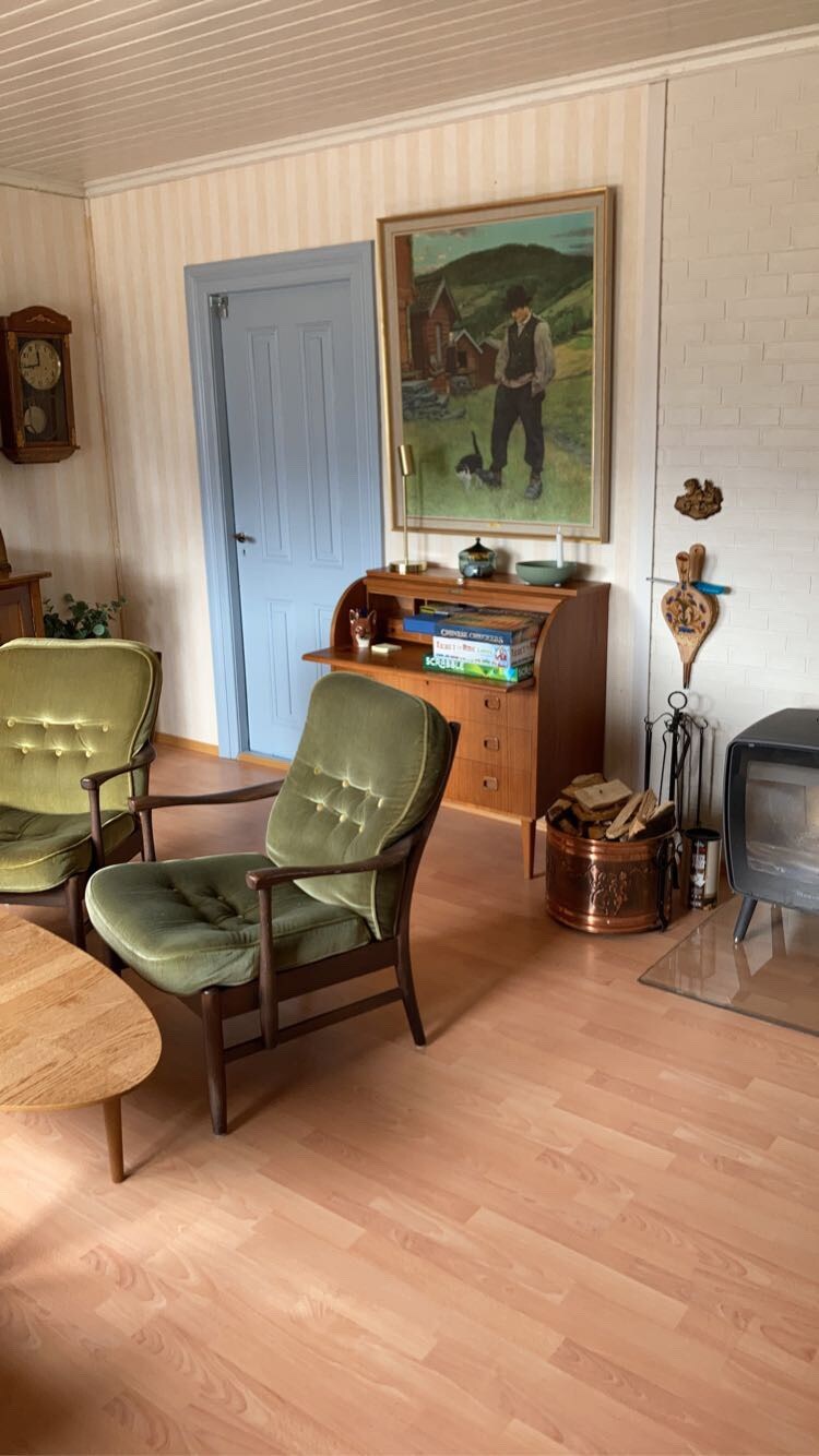 A cozy sitting area features two green chairs positioned around a wooden table. A painting depicts a rural scene on the wall, accompanied by a wooden bookshelf displaying various books and decorative items. A small, metal stove adds warmth to the space.
