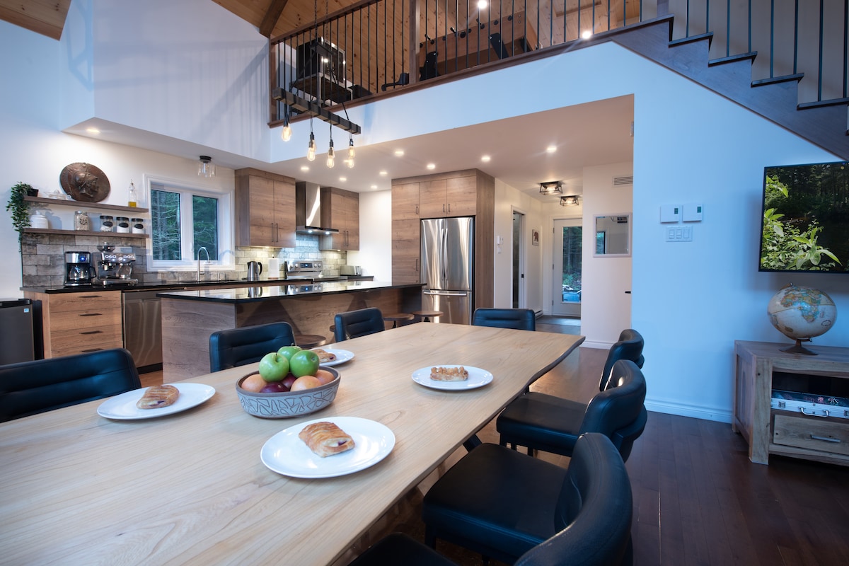 A spacious dining area is highlighted by a large wooden table with several plates arranged on it. The kitchen is visible in the background, showcasing modern appliances and cabinetry. Warm lighting complements the wood features, contributing to an inviting atmosphere.