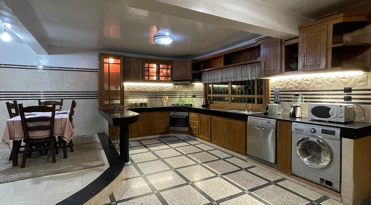 A spacious kitchen is equipped with wooden cabinets and modern appliances, including a washing machine and microwave. A dining table with four wooden chairs is situated nearby. Natural light enters through the window, enhancing the warm tones of the interior.