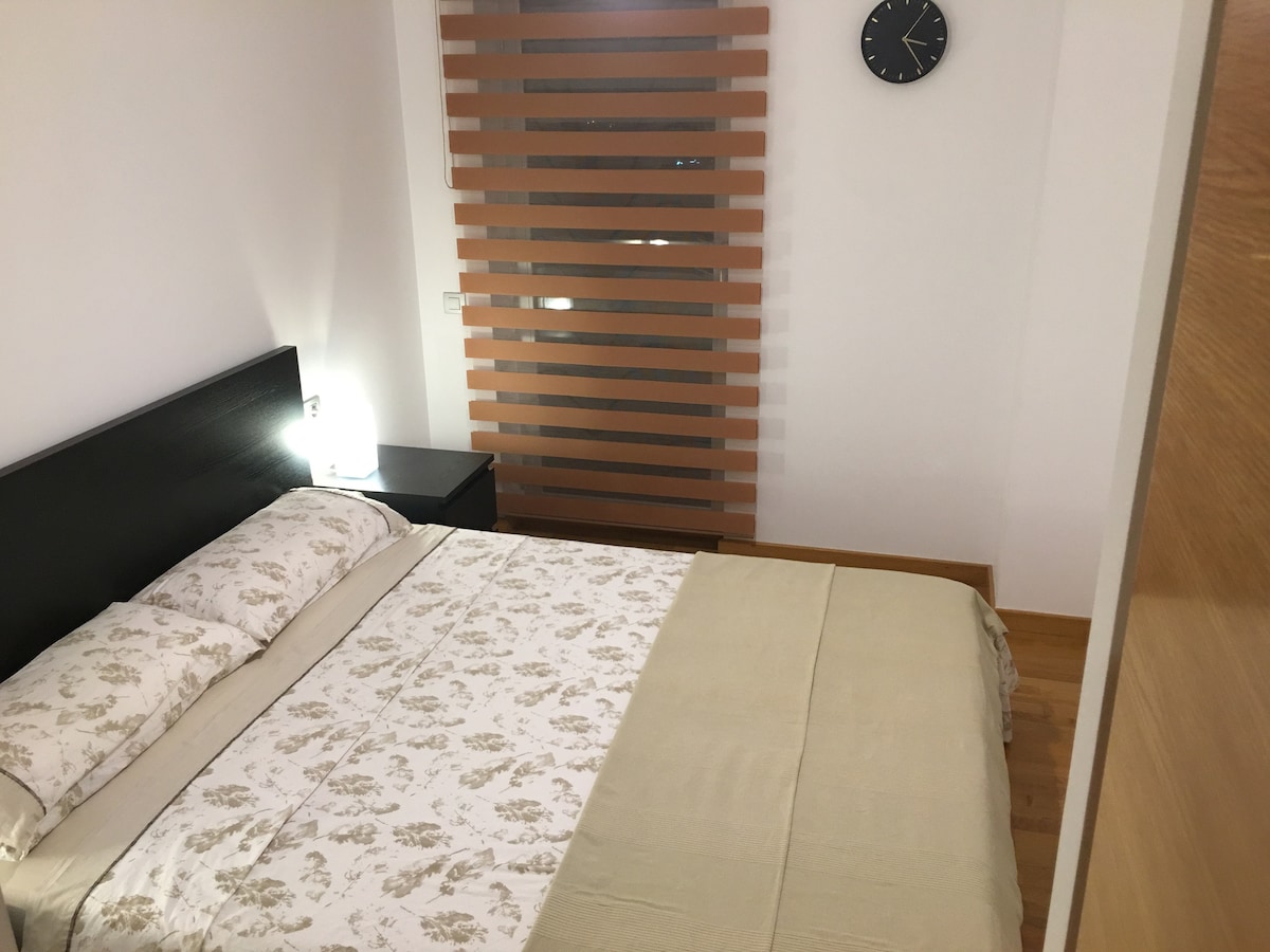 A private bedroom features a spacious bed with a floral-patterned duvet cover. A small bedside table with a lamp is positioned beside the bed. Wooden slats provide privacy while allowing light to filter in, and a clock is mounted on the wall.