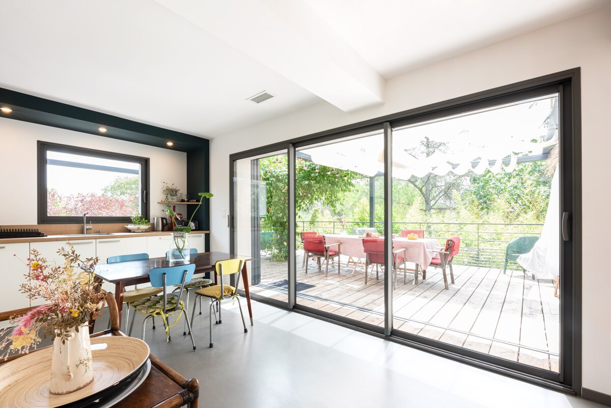 A spacious dining area is presented with a wooden table surrounded by colorful chairs. Floor-to-ceiling glass doors offer a view of a sunny terrace adorned with outdoor seating, extending towards a lush garden beyond.
