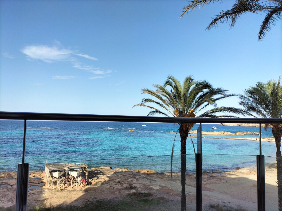 A balcony view showcases a tranquil shoreline with clear blue waters extending to the horizon. Palm trees frame the scene, while a rustic structure sits on the rocky beach, adding a natural element to the coastal landscape.