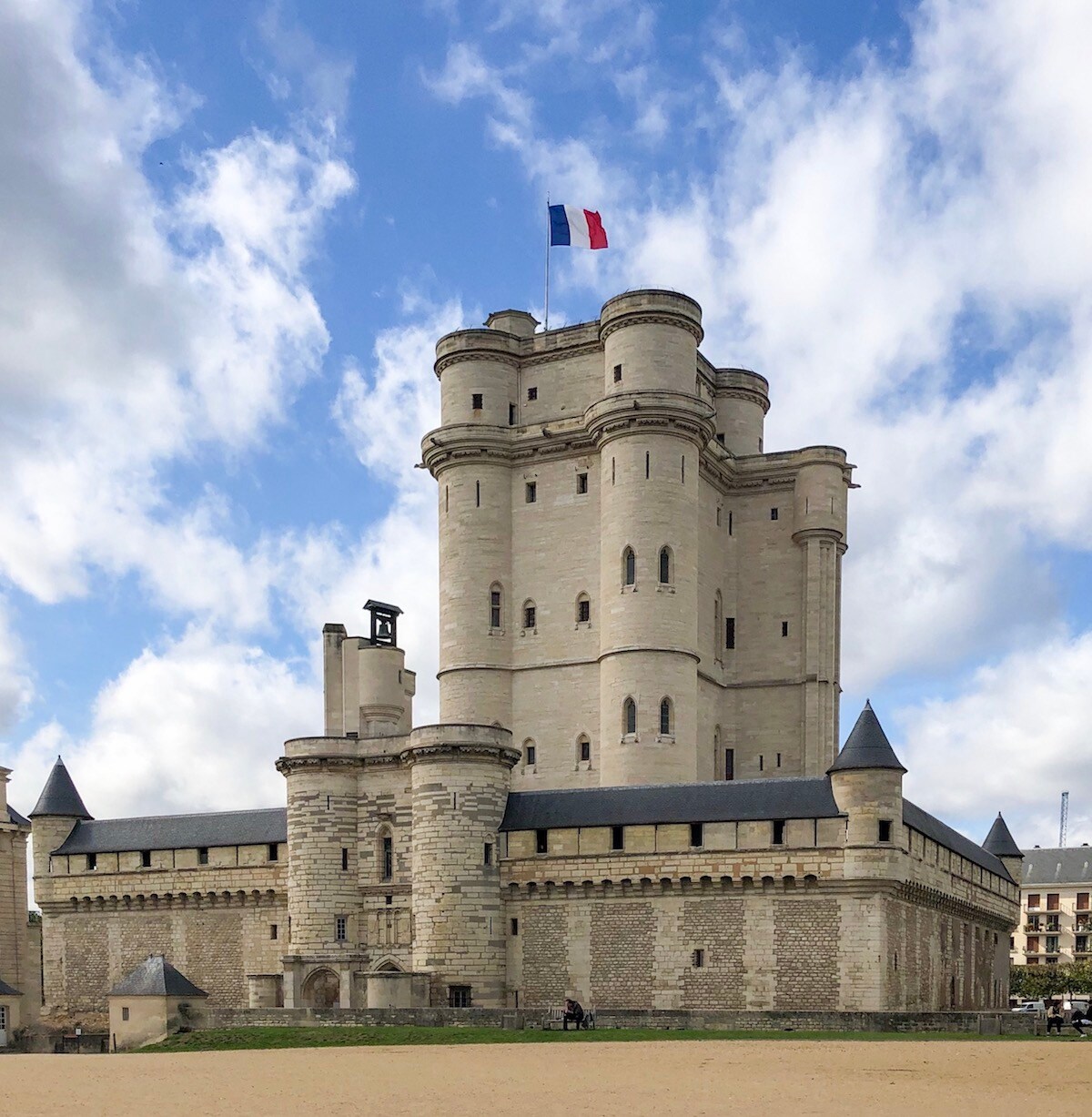 The imposing Château de Vincennes is depicted with its tall stone towers and distinctive cylindrical structures. A French flag flutters atop the main tower against a backdrop of clouds, showcasing the castle's historical architecture and sturdy fortifications.