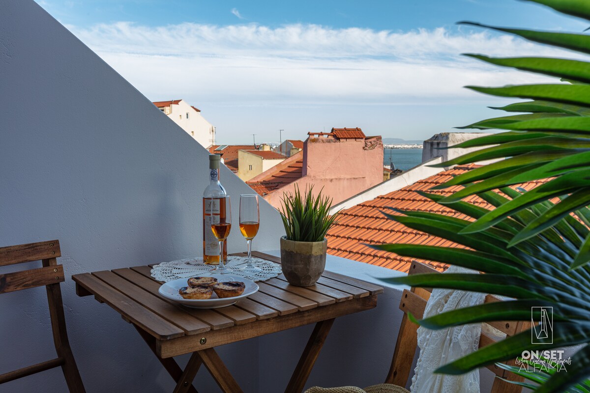 A small balcony features a wooden table set with two glasses of orange drink and a plate of cookies. A potted plant adds a touch of greenery, while terracotta rooftops and a clear blue sky create a serene backdrop.