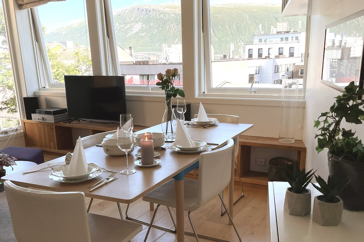 A well-appointed dining area features a light wood table set for four, with white plates and wine glasses arranged neatly. Natural light fills the space through large windows, offering views of the surrounding mountains. An elegant vase with flowers adds a touch of greenery.