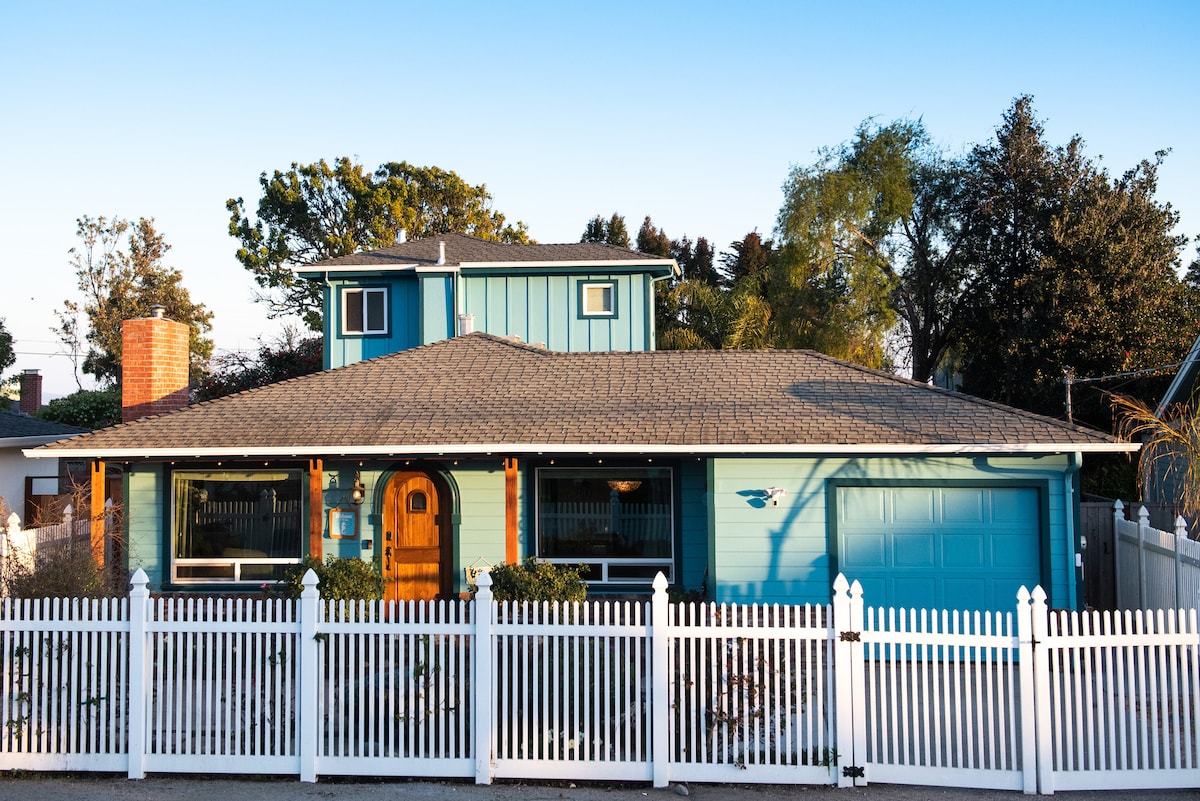 A light blue bungalow with a charming front entry features a wooden door and white picket fence. A well-kept lawn surrounds the home, and a cozy garage is visible on the right side. The setting is peaceful, with trees in the background under a clear blue sky.