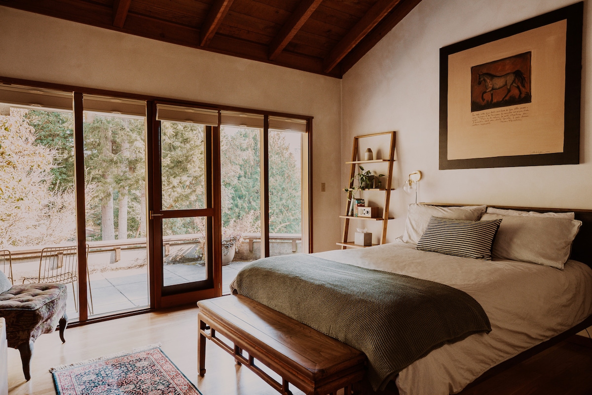 A spacious bedroom features a large bed with soft bedding. Natural light pours through sliding glass doors, which lead to a private patio. A serene artwork hangs above the bed, complemented by a cozy bench at the foot and a decorative rug on the wooden floor.
