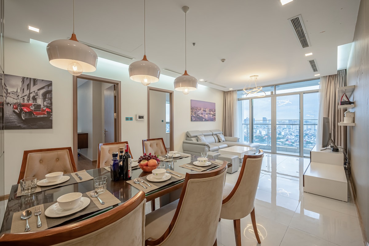 A modern dining area is illuminated by pendant lights, featuring a glass dining table set for six with plates and cutlery. A comfortable sofa is visible in the background, next to large windows offering a city view. Wall art adds a touch of character to the space.