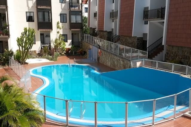 A community pool is set within the apartment complex, featuring a rounded shape with light blue water surrounded by a safety fence. Lush greenery adds a natural touch to the space, while the buildings provide a structured backdrop.