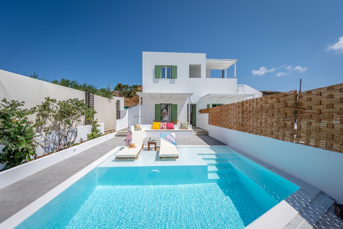 A sleek villa is showcased under a clear blue sky, featuring a private swimming pool surrounded by sun loungers. Bright cushions add color to the lounging area, while lush greenery provides a natural backdrop. A spacious terrace is accessible from the indoor living area.