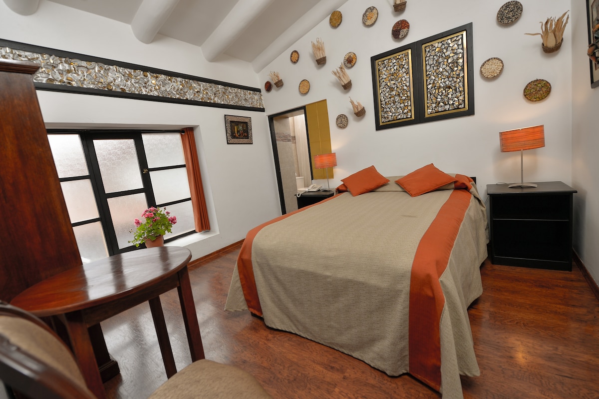 A cozy sleeping area features a spacious bed dressed in neutral tones with orange accents. Decorative plates adorn the walls, complemented by framed artwork. Natural light enters through the window, illuminating a small wooden table with two chairs in the corner.