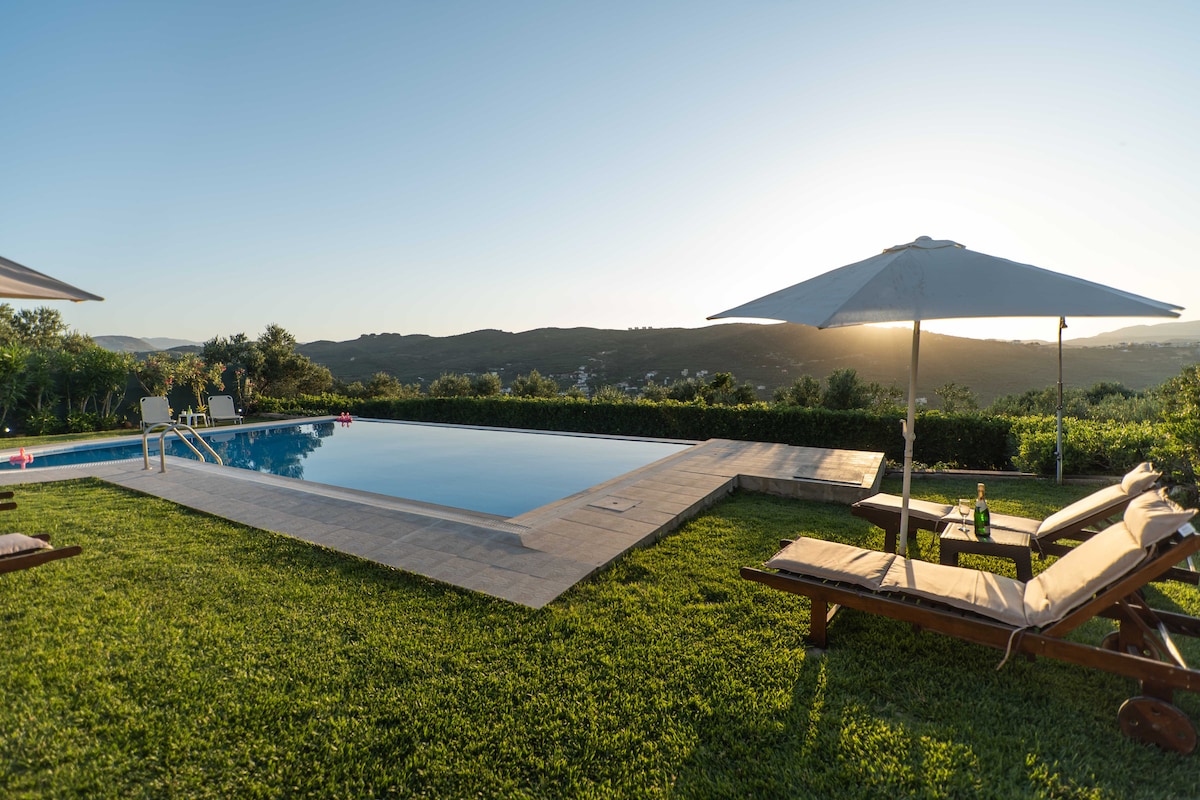 The image showcases a serene swimming pool surrounded by lush greenery, with sun loungers positioned nearby under umbrellas. The sun sets in the background, casting a warm glow over the landscape, while distant hills create a peaceful horizon.