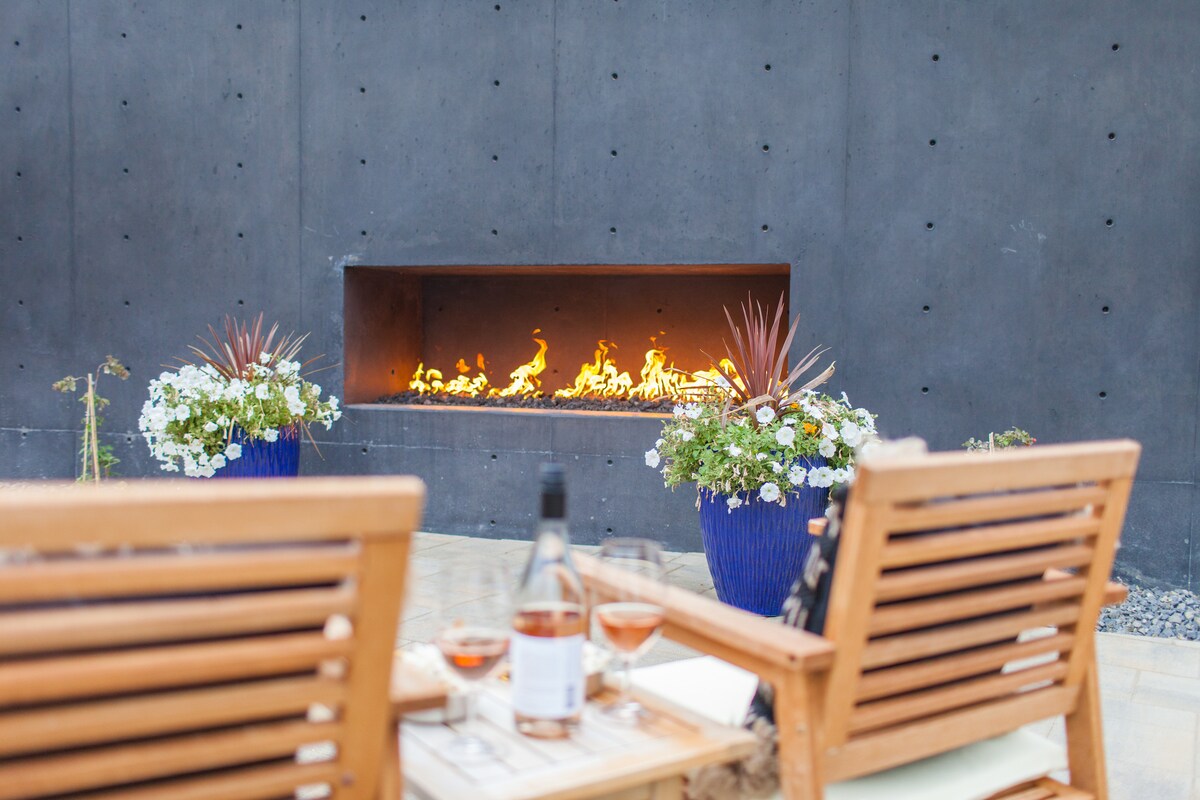 An outdoor seating area is featured, showcasing two wooden chairs positioned in front of a gas fire feature. Flanking the fire, vibrant flower pots with blooming plants add a touch of color to the space, creating an inviting atmosphere for relaxation.