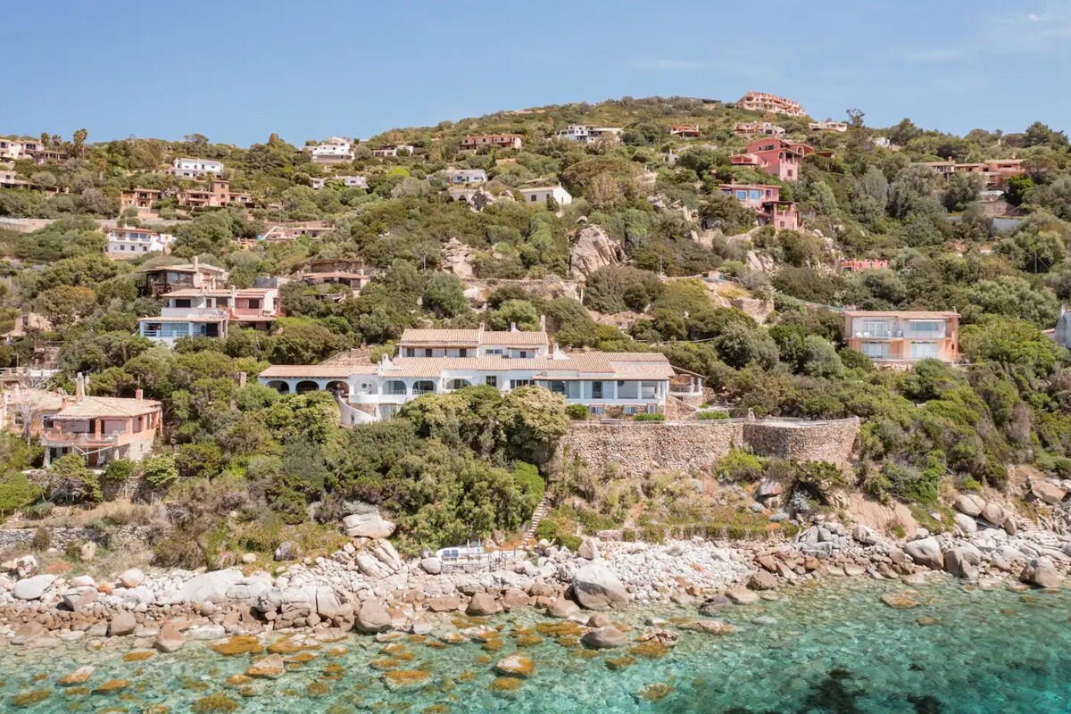 The villa's exterior is showcased, positioned alongside a clear turquoise sea with rocky shores. Lush greenery surrounds the property, while other villas are seen on the hillside in the background, adding to the tranquil setting.