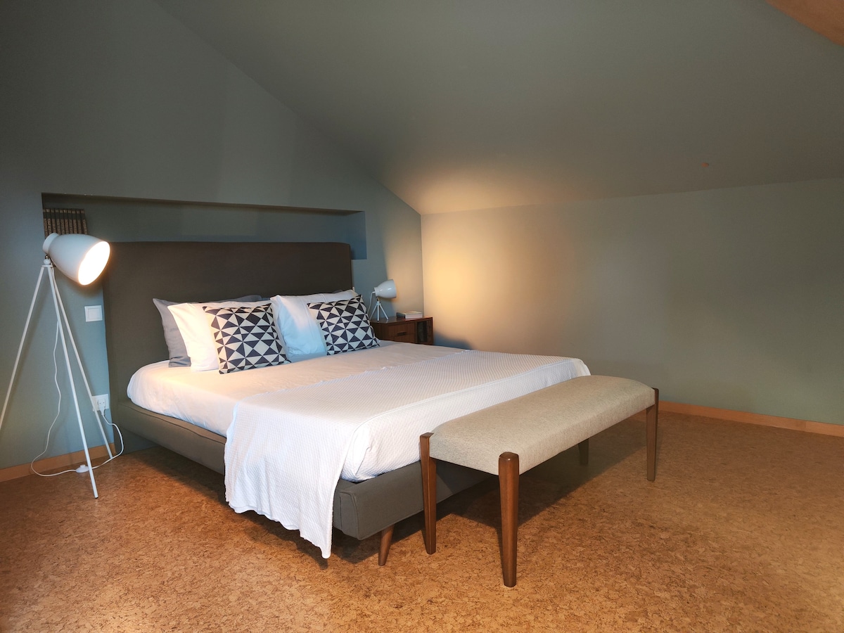 A spacious bedroom features a large bed with a light-colored bedspread and patterned throw pillows. Soft lighting from sleek lamps flanks the bed, illuminating the warm cork flooring. A minimalist bench rests at the foot of the bed, contributing to the room's serene atmosphere.