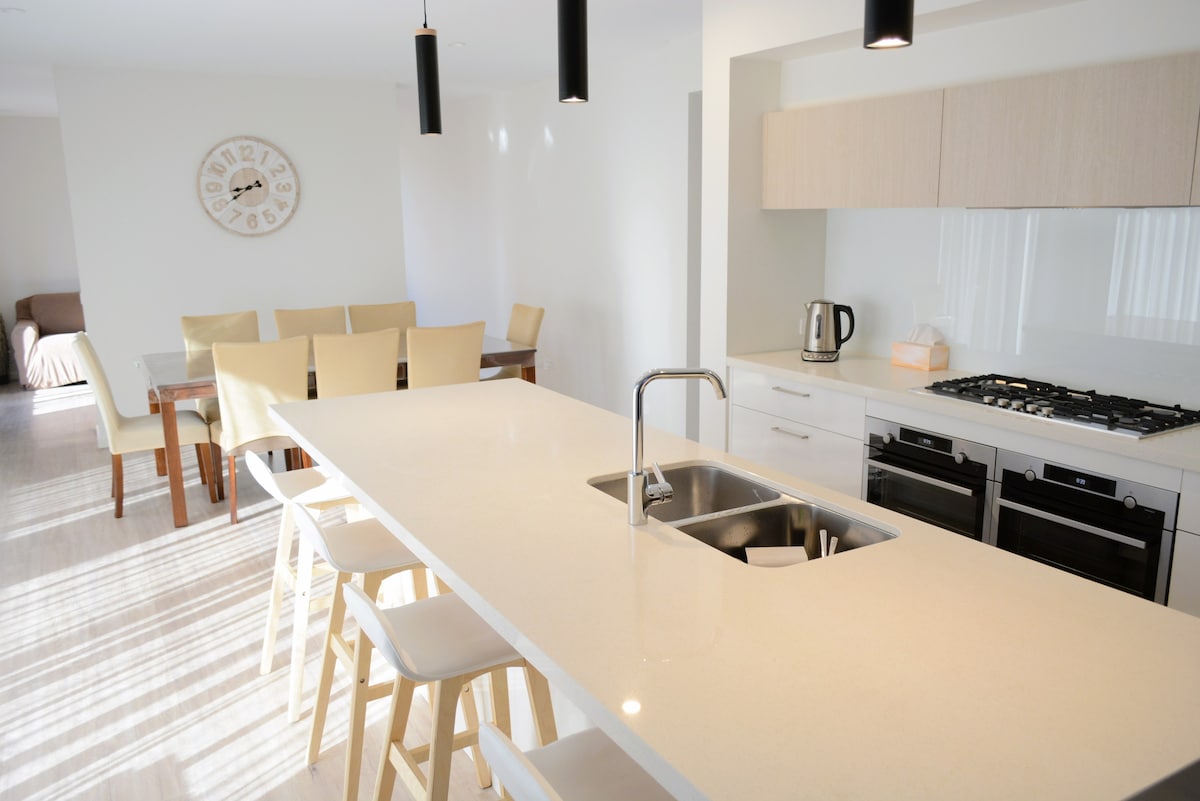 A modern kitchen features a light-colored stone countertop with a double sink and sleek stainless-steel appliances. Adjacent, a dining area is visible with a wooden table surrounded by light-colored chairs. Natural light filters through large windows, enhancing the open and airy design.
