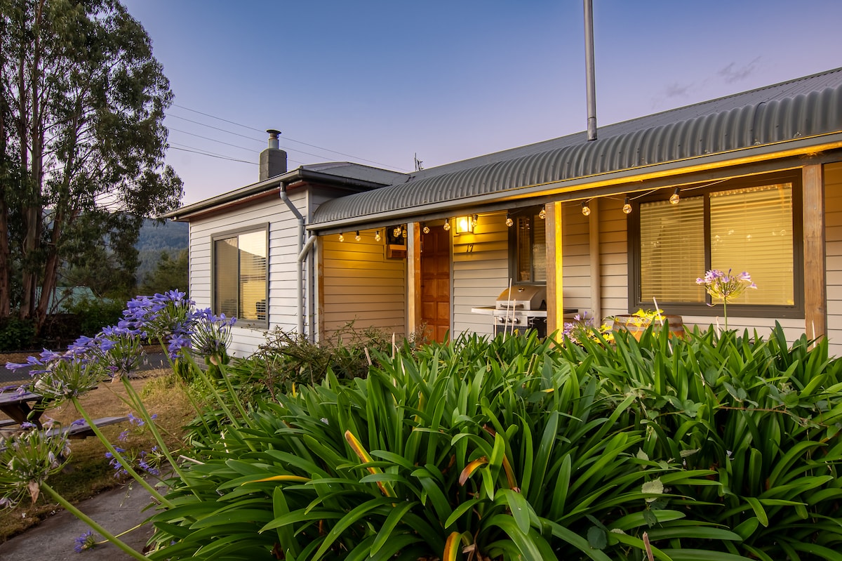 A charming home exterior is framed by lush green plants and flowers, with a welcoming entrance highlighted by warm outdoor lighting. Windows provide a glimpse into the interior, while trees and mountains create a serene backdrop.