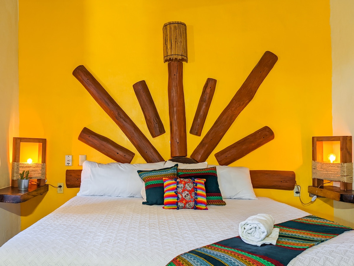 A spacious bed is showcased against a warm yellow wall, adorned with vibrant throw pillows. Wooden decor pieces fan out from a central lamp, providing a unique focal point. Subtle lighting from bedside lamps adds a calm ambiance to the room.