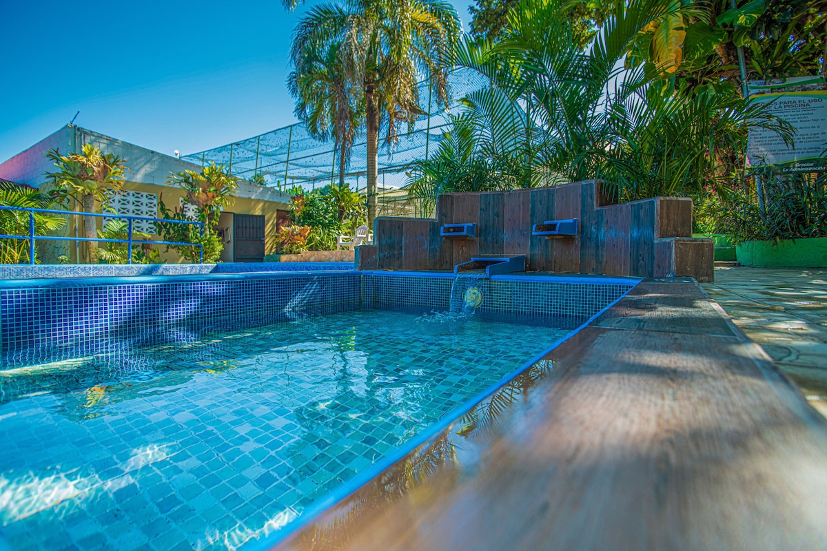 A tranquil pool area is surrounded by lush greenery, featuring a tiled pool with a gentle waterfall. Sunlight reflects off the water's surface, creating a serene atmosphere. In the background, a multi-purpose area is visible, with additional seating and plant life enhancing the setting.