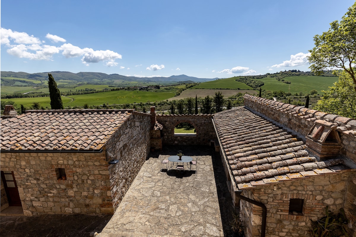A spacious outdoor area is visible, featuring stone-paved surfaces and a table set for dining. Surrounding landscapes reveal rolling hills and lush greenery, while distant mountains create a scenic backdrop under a clear blue sky.