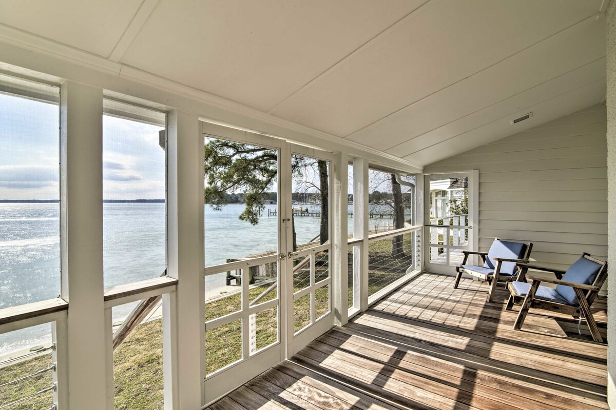 A screened-in porch overlooks the bay, featuring two blue lounge chairs positioned on a wooden deck. Large windows allow for unobstructed views of the water, enhancing the connection to the outdoor scenery. Sunlight streams in, creating a bright and open space.