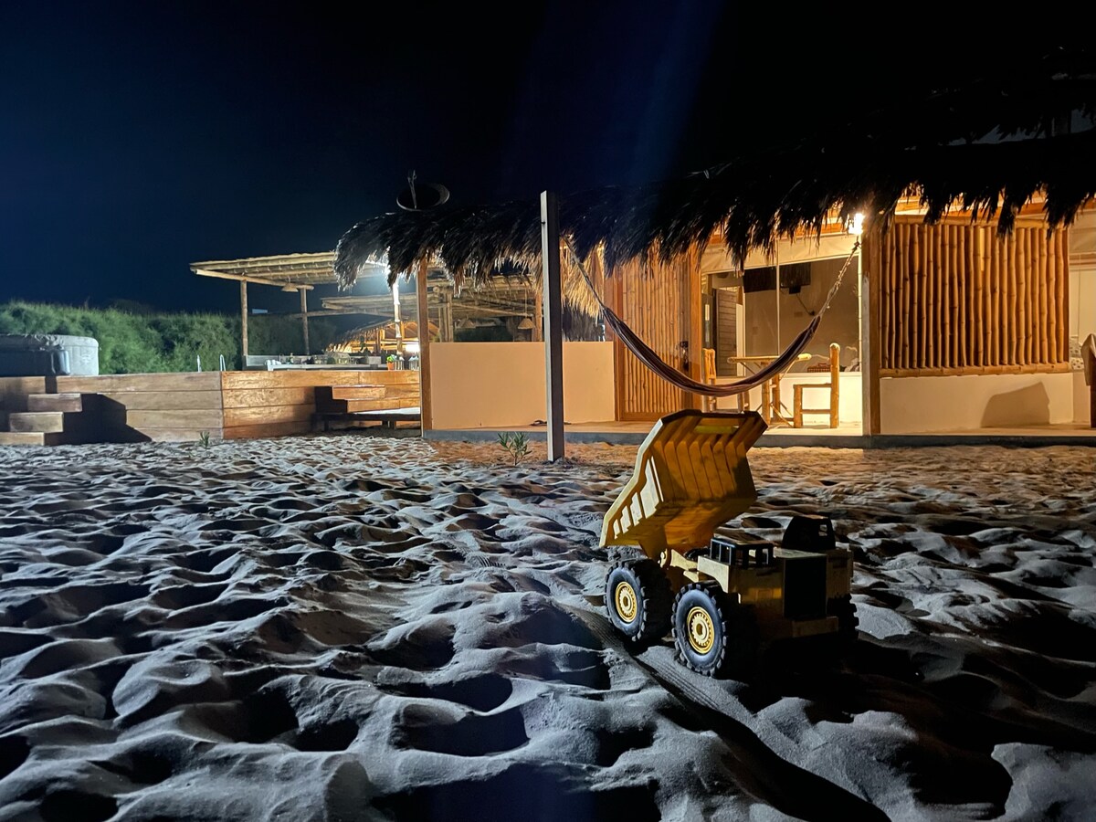 A toy dump truck is positioned on the sandy ground, illuminated by soft light from an outdoor lounge area. The gently lit structure features a thatched roof and a hammock, with the surrounding sand creating a tranquil beachfront environment at night.