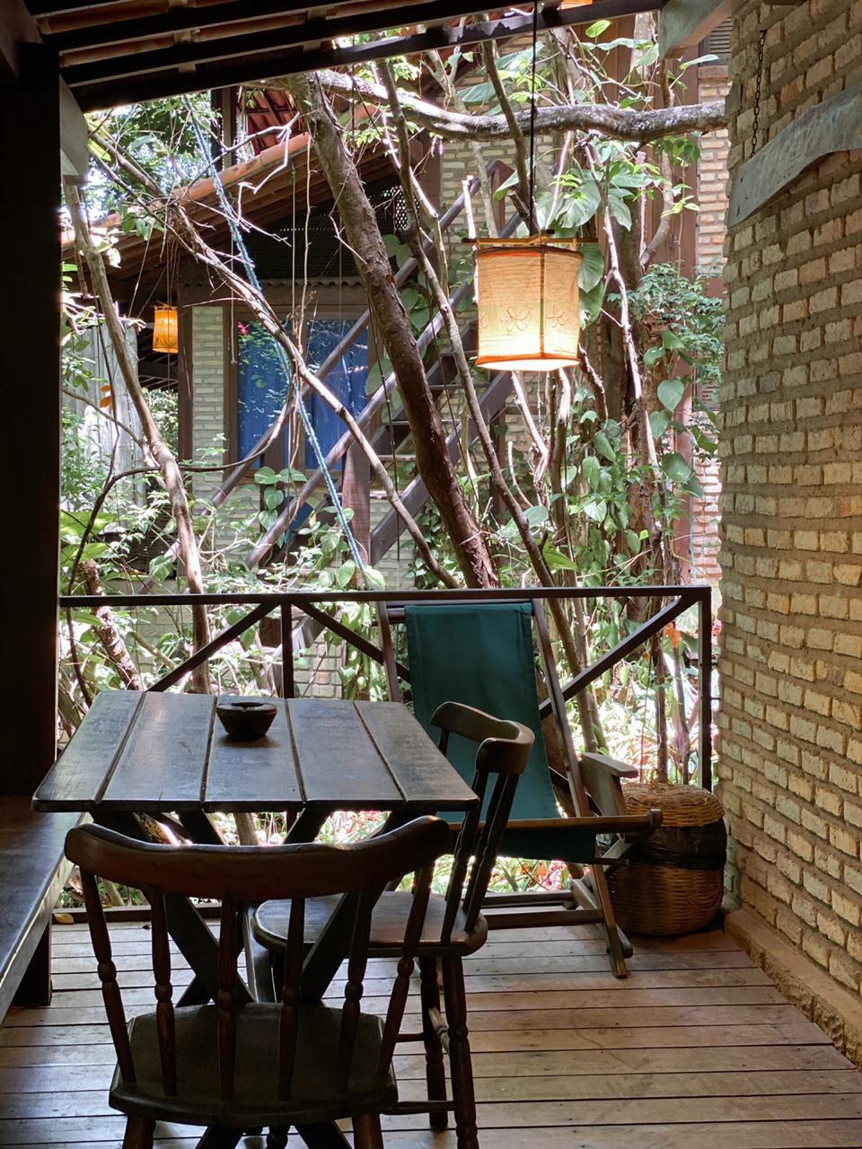 A wooden table is positioned on a wooden terrace, accompanied by a chair and a lounge chair. Lush greenery surrounds the area, with natural light filtering through the openings. A lantern hangs, providing a warm glow amid the vibrant foliage.