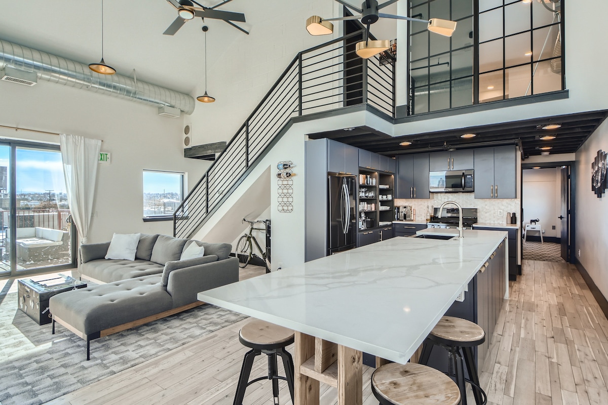 An open-concept living area showcases a modern kitchen with a large quartz island and sleek dark cabinetry, accompanied by high-end appliances. A spacious sectional sofa sits nearby, offering a comfortable seating option. Light filters in through large windows, enhancing the inviting atmosphere.