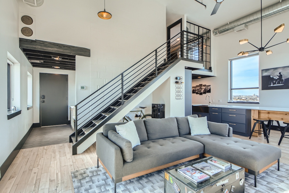 The open living area features a modern sectional sofa arranged on a patterned rug, with a staircase leading to the upper level. Large windows provide natural light, while a contemporary dining table and kitchen with dark cabinetry are visible in the background.