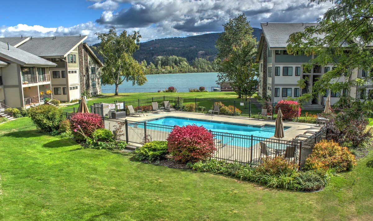 A landscaped outdoor area features a swimming pool surrounded by a secure fence, with lounge chairs positioned nearby. Colorful shrubs and greenery frame the pool, while a peaceful lake and mountains are visible in the background under a partly cloudy sky.