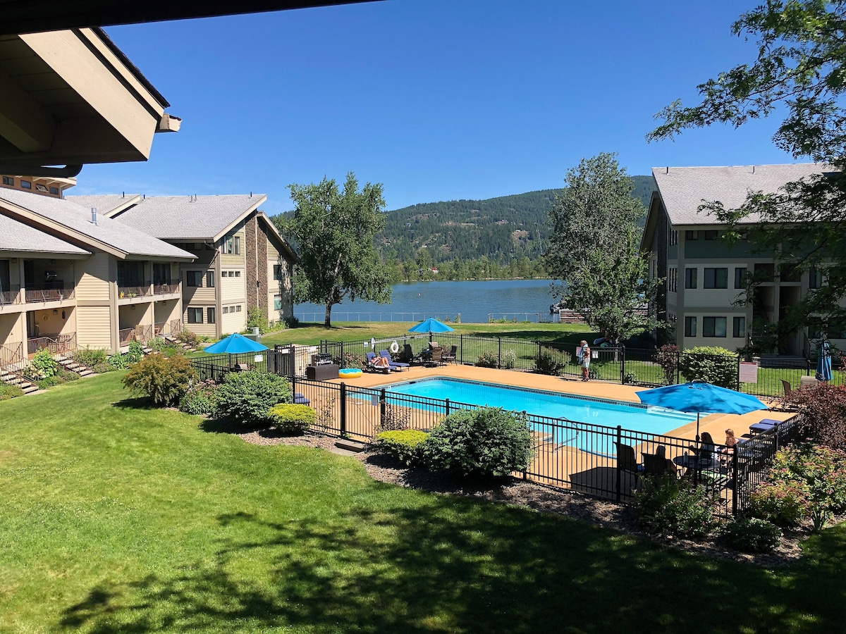 The image captures a sunny outdoor pool area surrounded by well-maintained lawns and gardens. Bright blue lounge chairs are positioned by the pool, with a view of the lake and distant mountains providing a scenic backdrop. The clear sky contributes to a vibrant atmosphere.