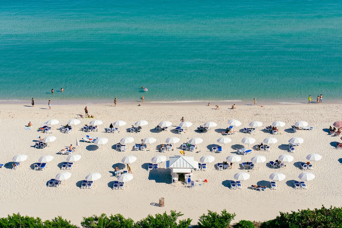 An aerial view of a sandy beach featuring rows of lounge chairs topped with white umbrellas. The calm turquoise water gently meets the shore, with visitors enjoying the beach. A small beach hut is positioned centrally, surrounded by greenery.