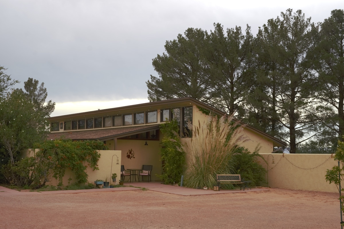 The exterior of a welcoming home showcases warm earth-toned walls and large windows. Lush greenery and ornamental plants surround a small outdoor seating area, while a landscaped pathway leads to the entrance. The architecture reflects a harmonious blend with the natural surroundings.