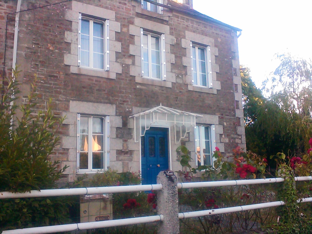 The exterior of a stone house is visible, showcasing unique architectural features, including multiple windows adorned with shutters. A blue front door is framed by a white canopy. Lush greenery and blooming flowers enhance the inviting appearance of the home.